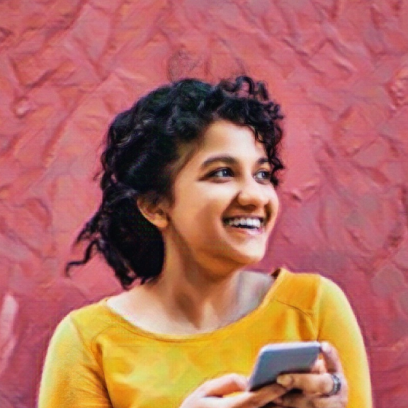 Young person in front of a red wall, wearing a yellow shirt is smiling and holding a cell phone
