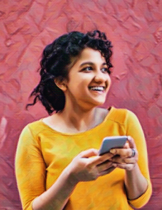Young person in front of a red wall, wearing a yellow shirt is smiling and holding a cell phone