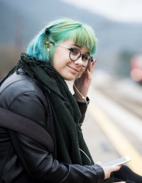 A content looking teenager with blue green hair sits by a train track